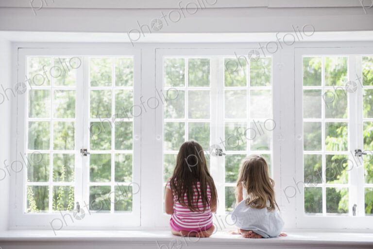 Two girls sitting side by side by a large window.