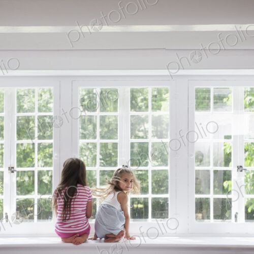 Two girls sitting side by side by a large window.