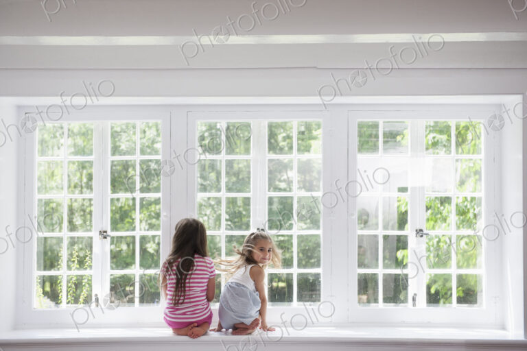 Two girls sitting side by side by a large window.