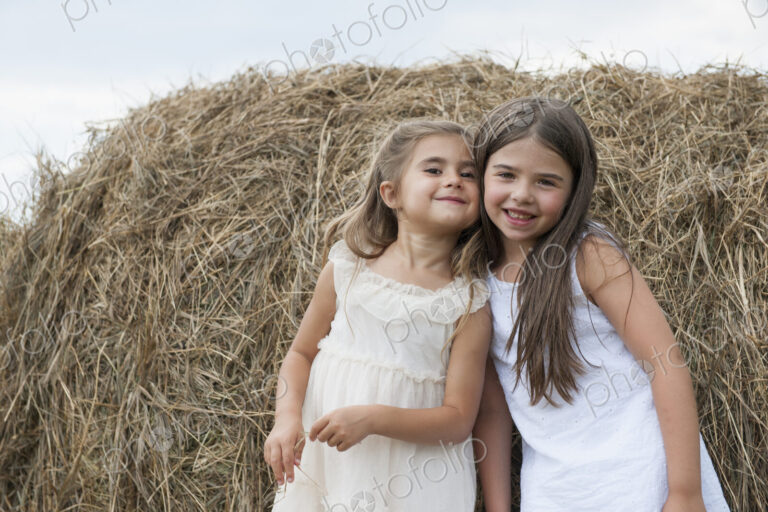 Two girls standing by a large haybale, smiling and sharing a secret.