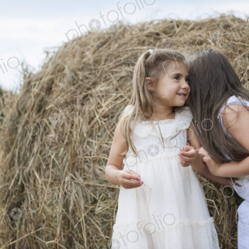 Two girls standing by a large haybale, smiling and sharing a secret. Two girls standing by a large haybale, smiling and sharing a secret.