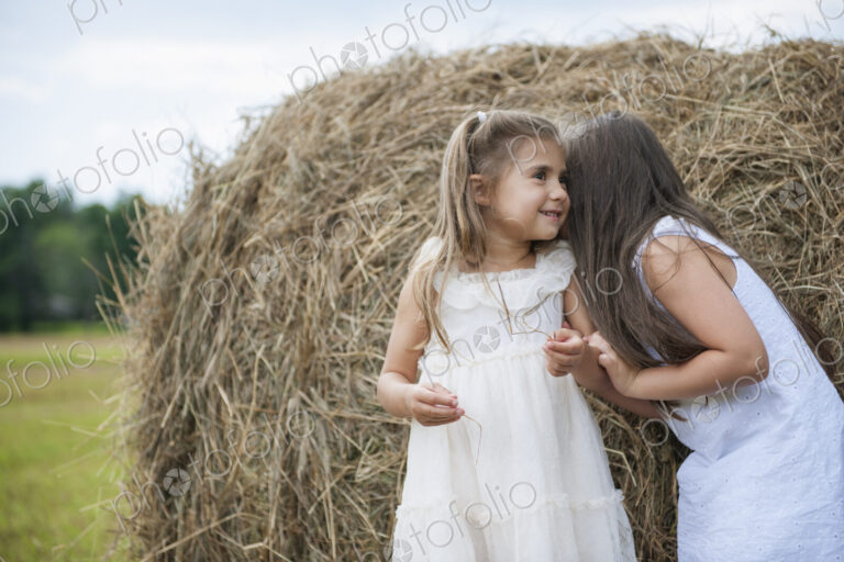 Two girls standing by a large haybale, smiling and sharing a secret.