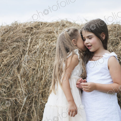 Two girls standing by a large haybale, smiling and sharing a secret. Two girls standing by a large haybale, smiling and sharing a secret.