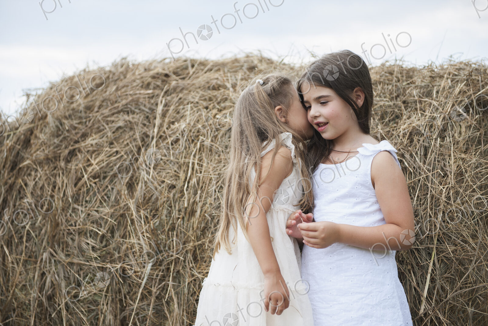 Two girls standing by a large haybale, smiling and sharing a secret.