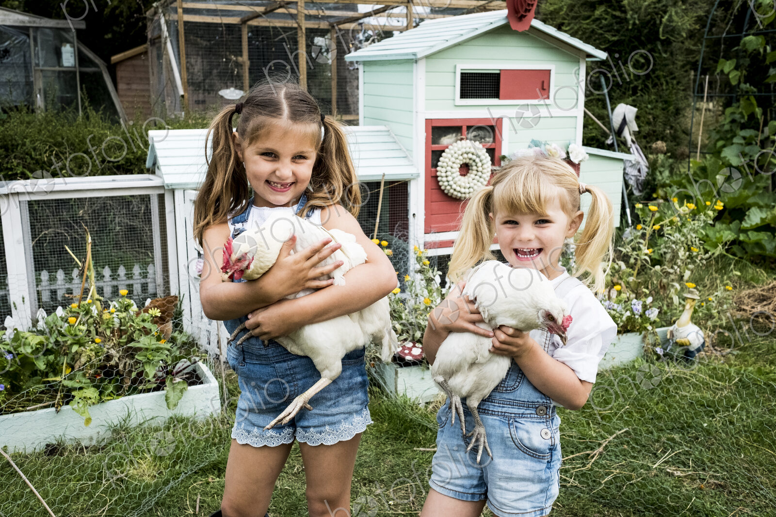 Two girls standing in front of hen house in a garden, holding white chickens, smiling…