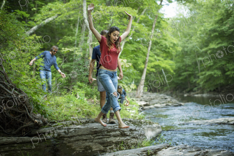 Two men and two women on the riverbank, one leaping across stepping stones.