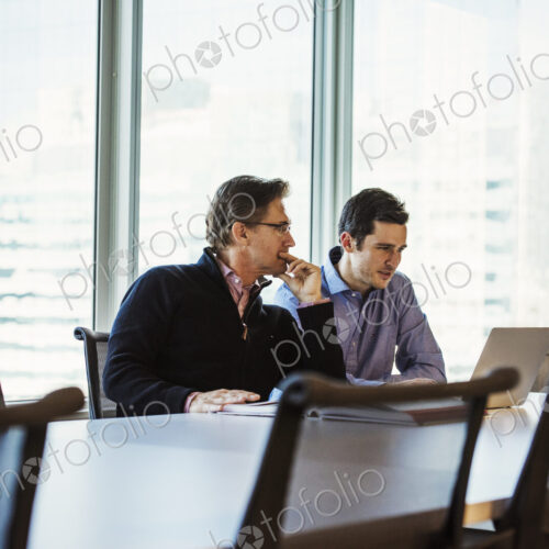 Two men at a table in a meeting room looking at a laptop computer.