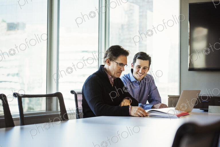 Two men at a table in a meeting room looking at a laptop computer.