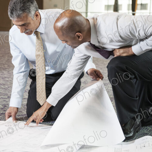Two men going over plans for a new office space while kneeling on an office carpet. Two men going over plans for a new office space while kneeling on an office carpet.