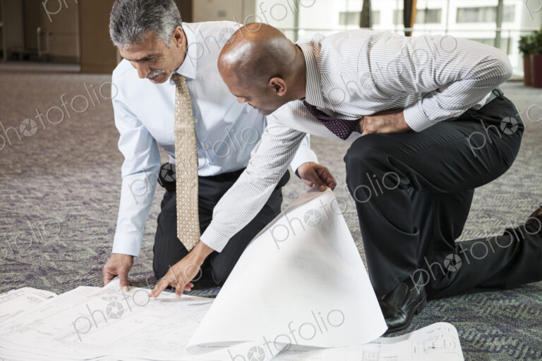 Two men going over plans for a new office space while kneeling on an office carpet.