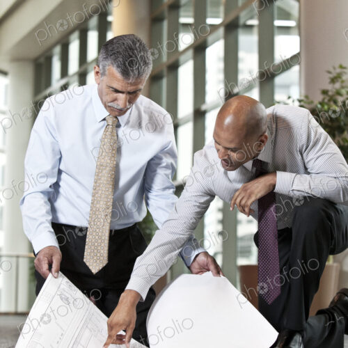 Two men going over plans for a new office space while kneeling on an office carpet. Two men going over plans for a new office space while kneeling on an office carpet.