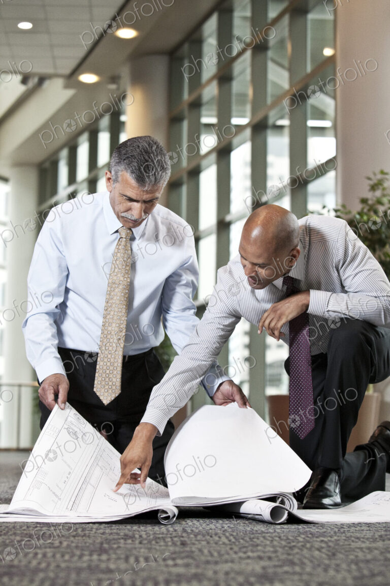 Two men going over plans for a new office space while kneeling on an office carpet.