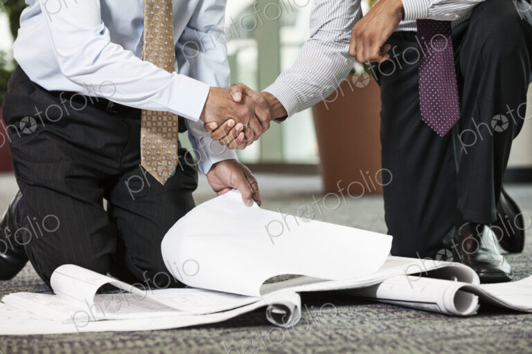 Two men going over plans for a new office space while kneeling on an office carpet.