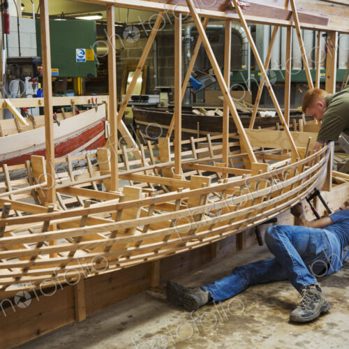 Two men in a boat-builder's workshop, working together on a wooden boat hull.
