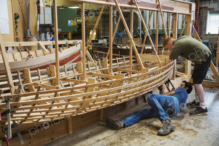Two men in a boat-builder's workshop, working together on a wooden boat hull.