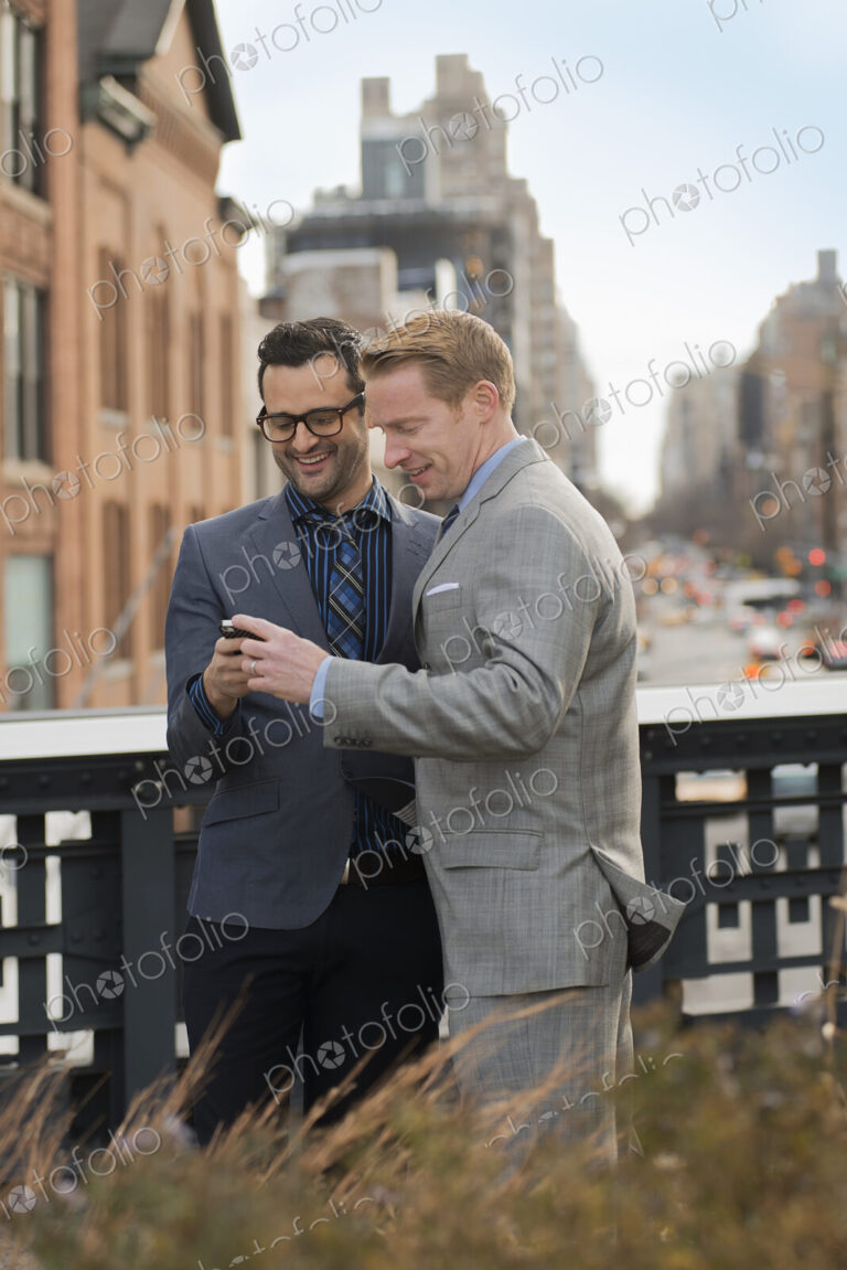 Two men in formal business clothes, standing side by side, looking at a cell phone…