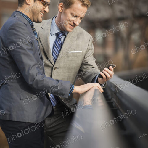 Two men in formal business clothes, standing side by side, looking at a cell phone… Two men in formal business clothes, standing side by side, looking at a cell phone…