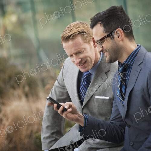 Two men in formal business clothes, standing side by side, looking at a cell phone… Two men in formal business clothes, standing side by side, looking at a cell phone…