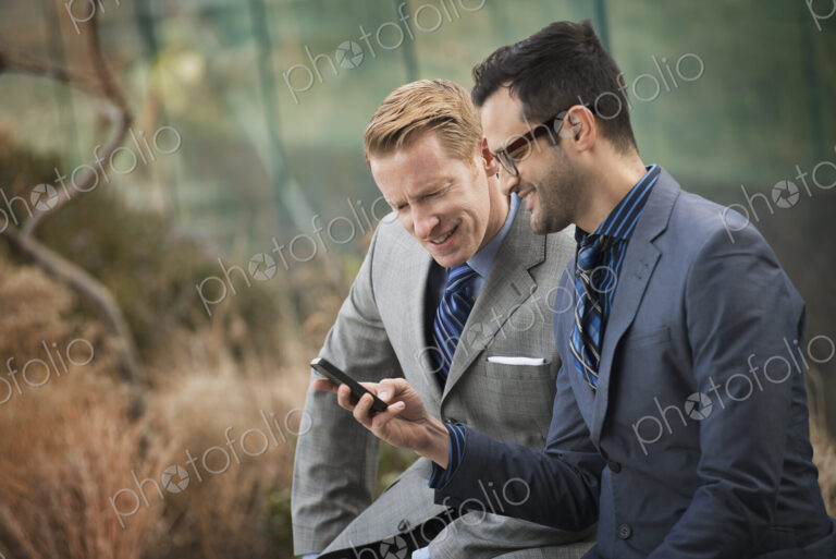 Two men in formal business clothes, standing side by side, looking at a cell phone…