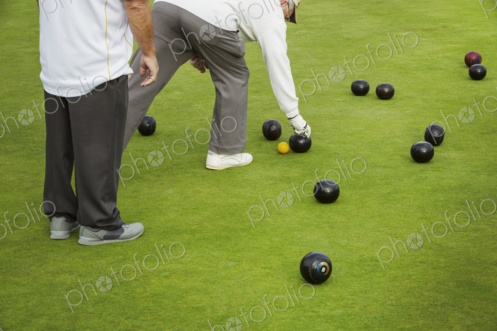 Two men on a bowls green, lawn bowls players, one bending down to pick up a wooden…