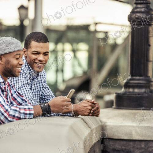 Two men side by side leaning on a parapet by a bridge looking at a smart phone