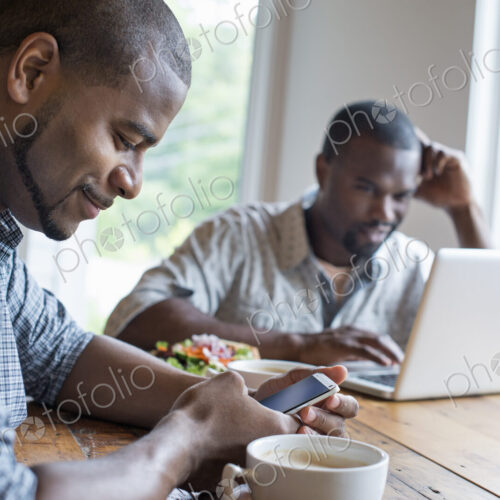 Two men sitting in a cafe. Using a laptop and a smart phone.
