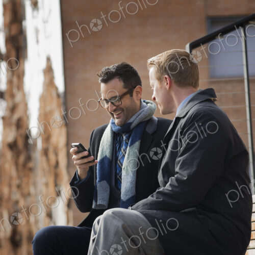 Two men sitting on a bench outside a large building in the city. One checking his… Two men sitting on a bench outside a large building in the city. One checking his…