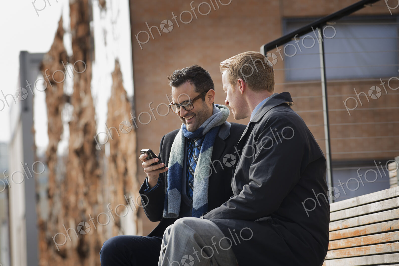 Two men sitting on a bench outside a large building in the city. One checking his…