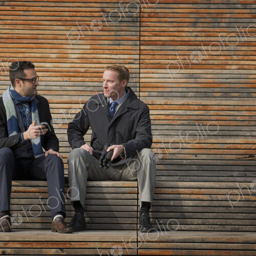 Two men sitting on a bench outside a large building in the city. One checking his… Two men sitting on a bench outside a large building in the city. One checking his…