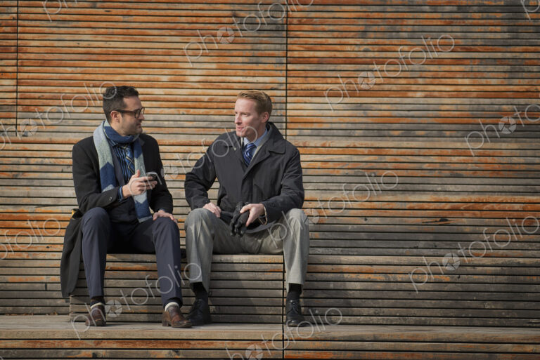 Two men sitting on a bench outside a large building in the city. One checking his…