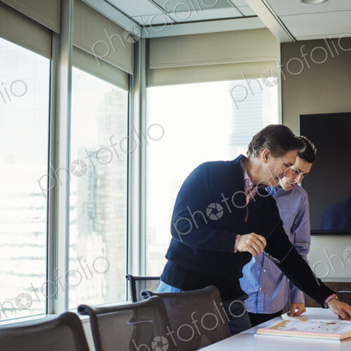 Two men standing at a table in a meeting room looking at an open book together.