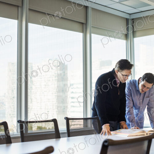 Two men standing at a table in a meeting room looking down at an open book together.
