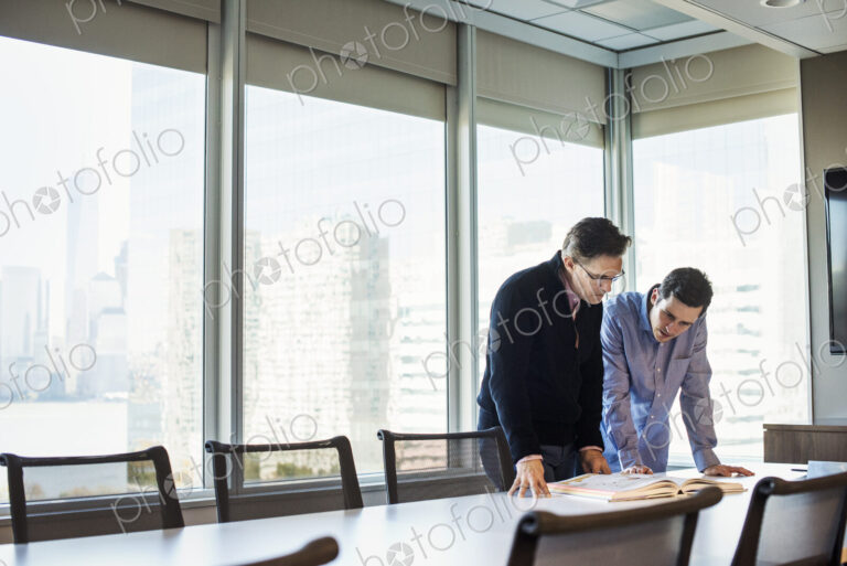 Two men standing at a table in a meeting room looking down at an open book together.