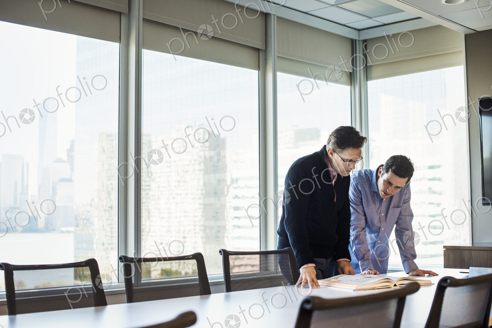Two men standing at a table in a meeting room looking down at an open book together.