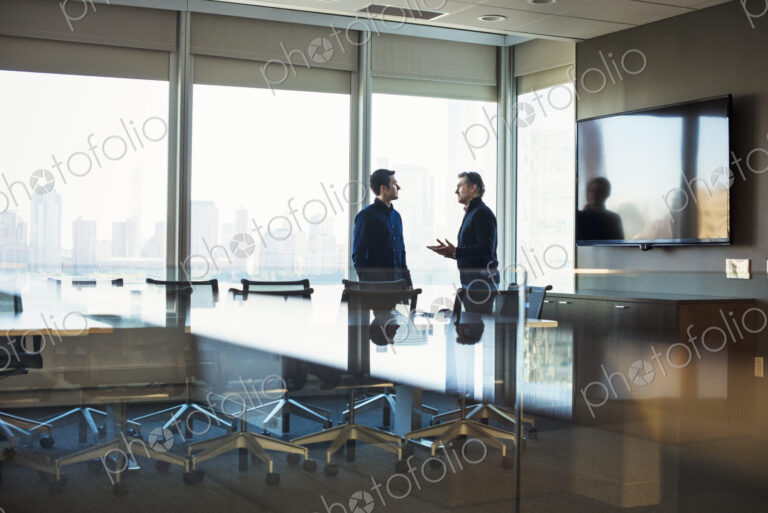Two men standing at a table in a meeting room talking to each other.