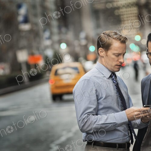 Two men standing together looking at a cell phone display on a busy street at dusk. Two men standing together looking at a cell phone display on a busy street at dusk.