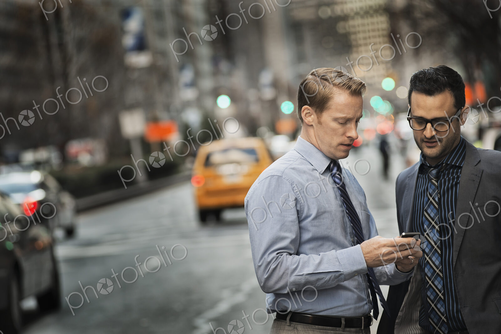 Two men standing together looking at a cell phone display on a busy street at dusk.