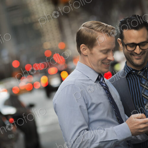 Two men standing together looking at a cell phone display on a busy street at dusk. Two men standing together looking at a cell phone display on a busy street at dusk.