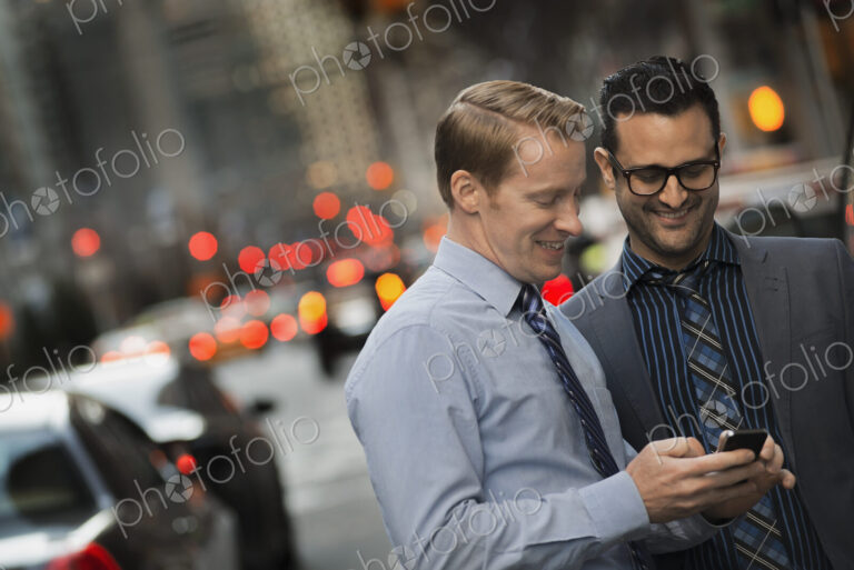 Two men standing together looking at a cell phone display on a busy street at dusk.