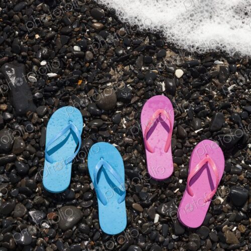 Two pairs of wet flip flops, one blue and one pink, rest on a rocky shoreline in…