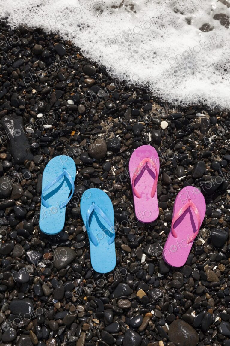 Two pairs of wet flip flops, one blue and one pink, rest on a rocky shoreline in…