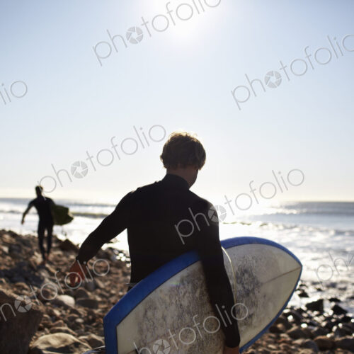 Two people in wetsuits on a beach, carrying their surfboards. Two people in wetsuits on a beach, carrying their surfboards.