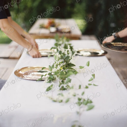 Two people leaning over a table laid outside with a white cloth and a central foliage…