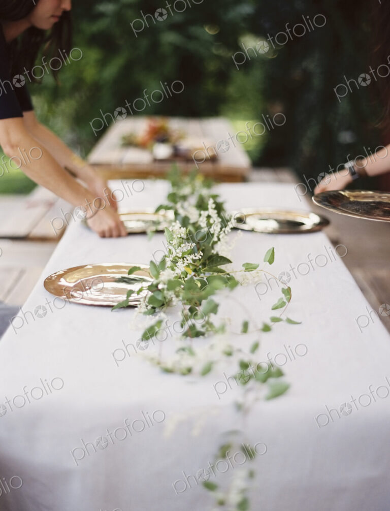 Two people leaning over a table laid outside with a white cloth and a central foliage…