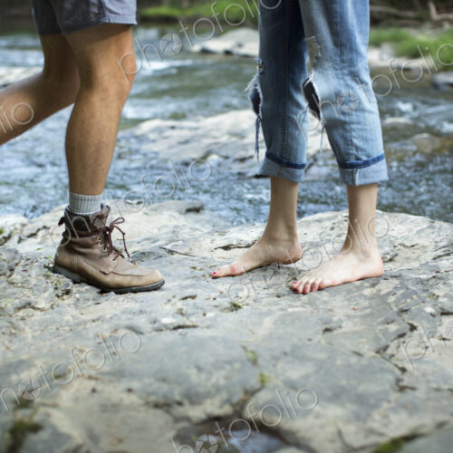 Two people on the rocks by a rushing river, man and woman, lower legs and feet.