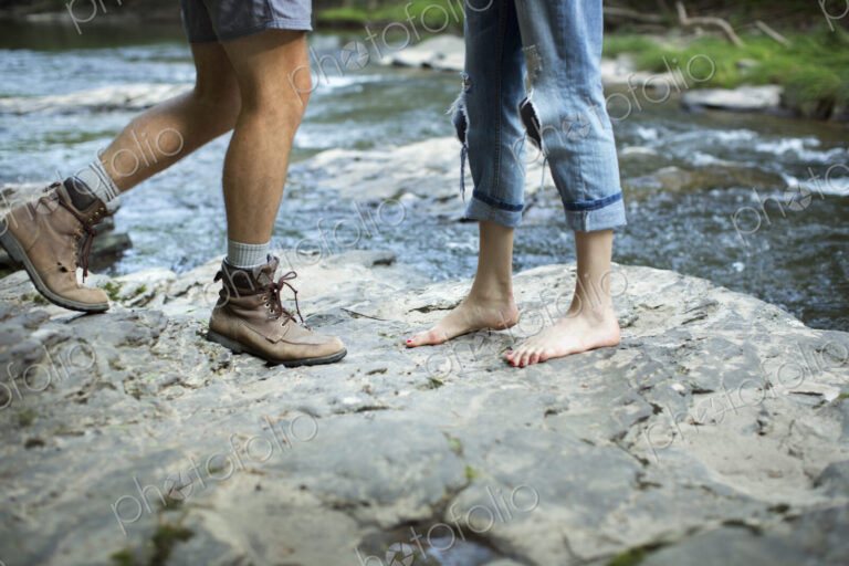 Two people on the rocks by a rushing river, man and woman, lower legs and feet.