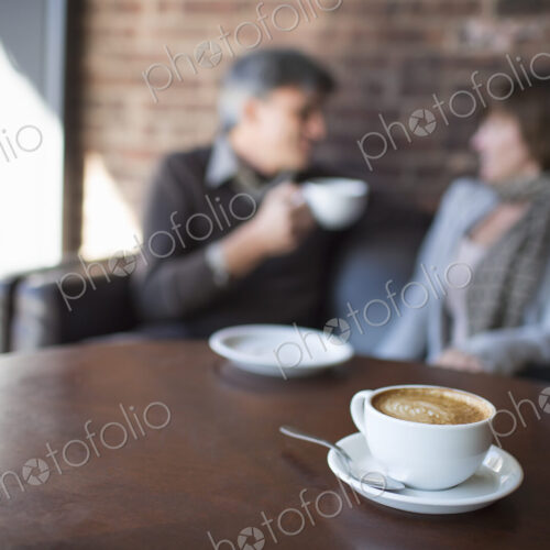 Two people sitting in a coffee shop. A man holding a white china of and drinking.…