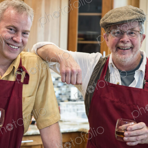 Two senior male friends have a drink before dinner in a home kitchen.