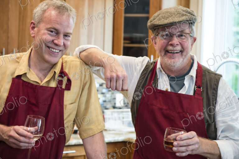Two senior male friends have a drink before dinner in a home kitchen.