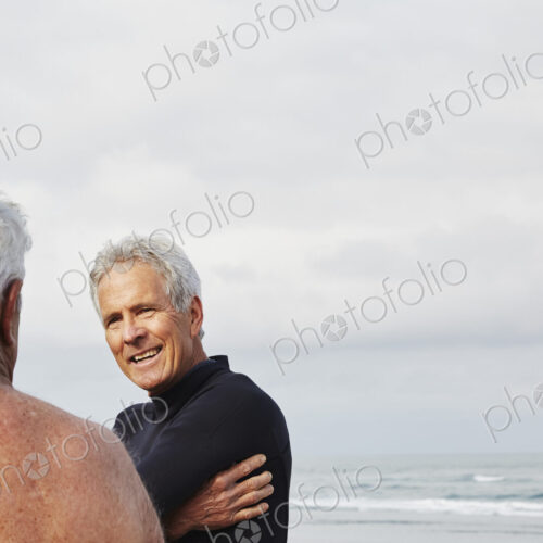 Two senior men standing on a beach chatting, one wearing a wetsuit. Two senior men standing on a beach chatting, one wearing a wetsuit.